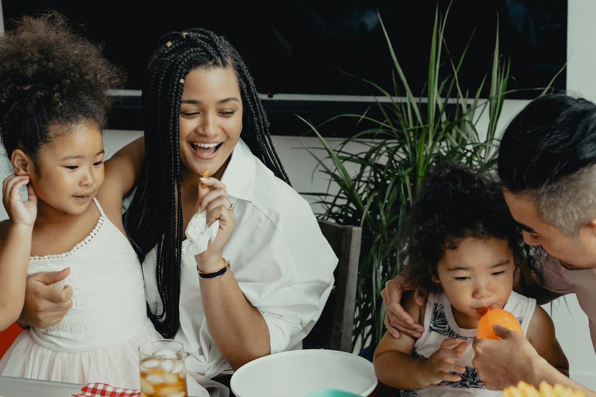 A family laughs together at a kitchen table: a mother sits with an older girl while a father helps a toddler bite into an orange slice.