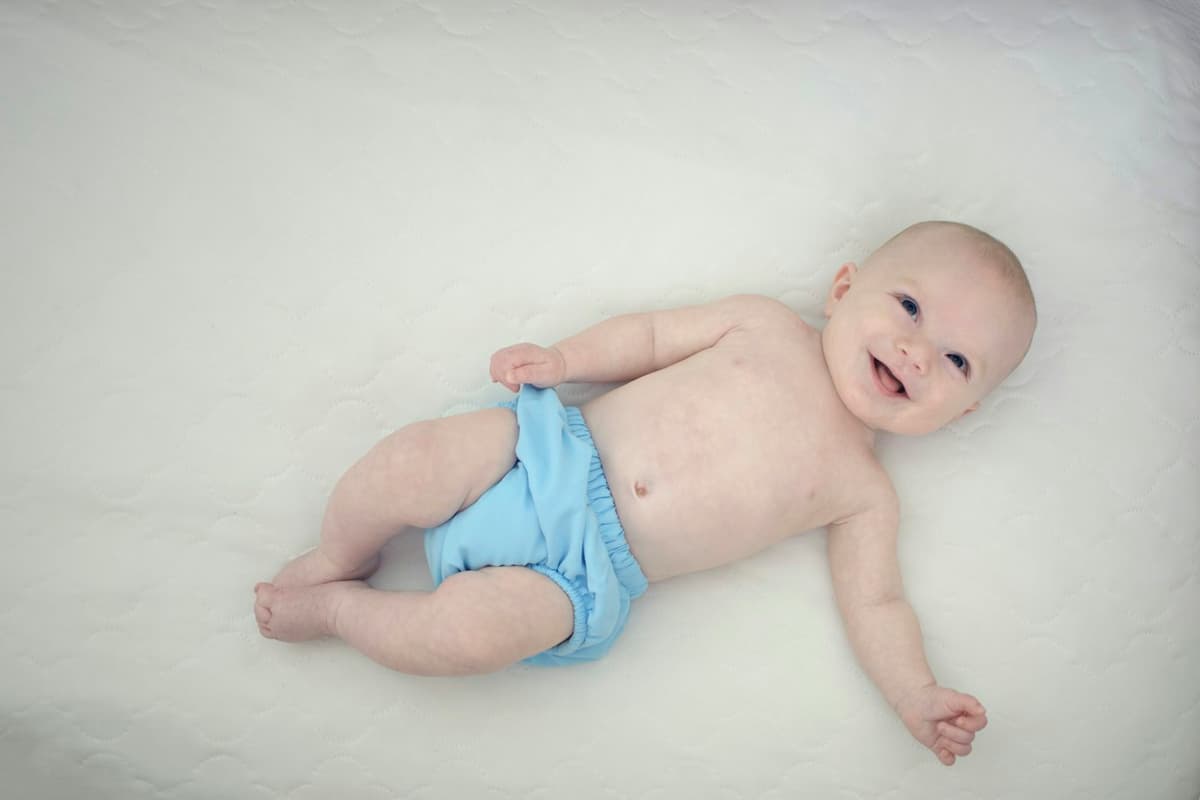 A smiling baby in a blue cloth diaper lies on a white quilted mattress.