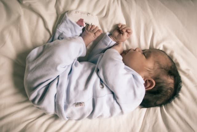 Um bebê dorme de lado sobre um lençol claro, de macacão azul-claro, com as mãos perto do rosto.