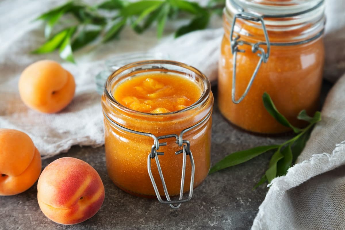 Two clip-top glass jars of apricot purée on a grey stone surface, with whole apricots and a sprig of green leaves beside them.