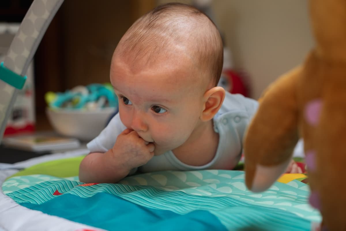 Um bebê de bruços num tapete de atividades, apoiado nos antebraços, com a cabeça erguida e uma das mãos na boca.