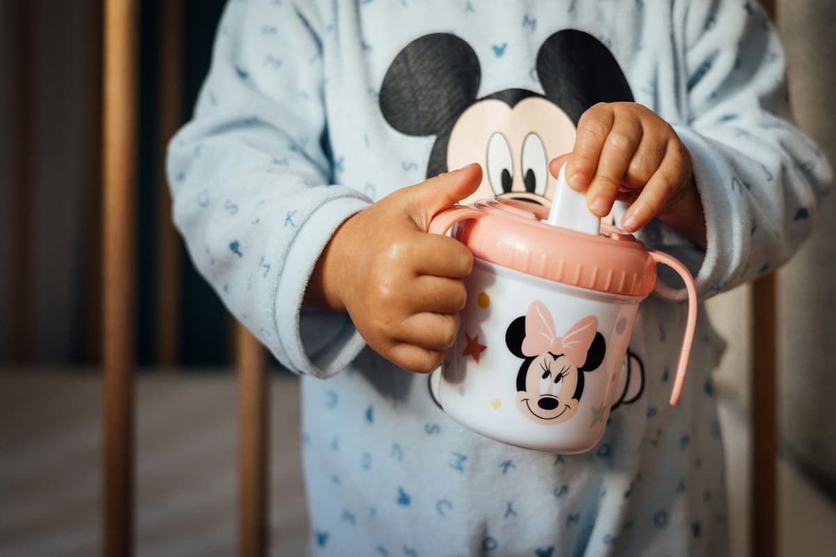 A toddler in printed pyjamas holds a pink and white spouted sippy cup with both hands.