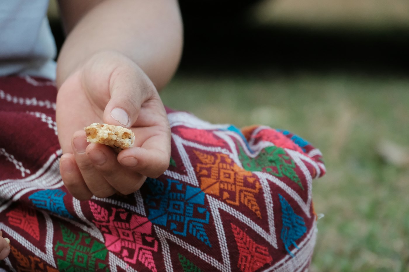 A child's hand holding a small piece of a cereal bar between thumb and fingers, above a brightly embroidered red blanket.
